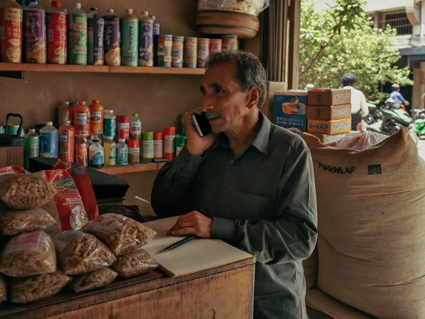 Shopkeeper on the phone beside a ledger notebook at the counter of a small neighbourhood grocery shop