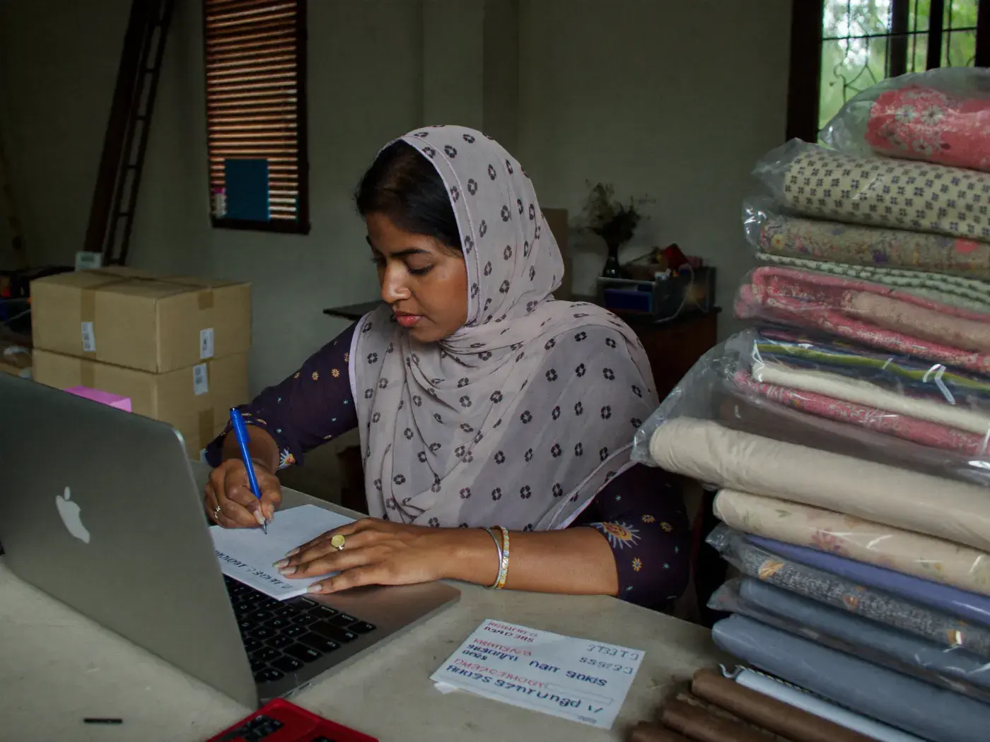 Small business owner writing a delivery label at a table stacked with parcel boxes and fabric